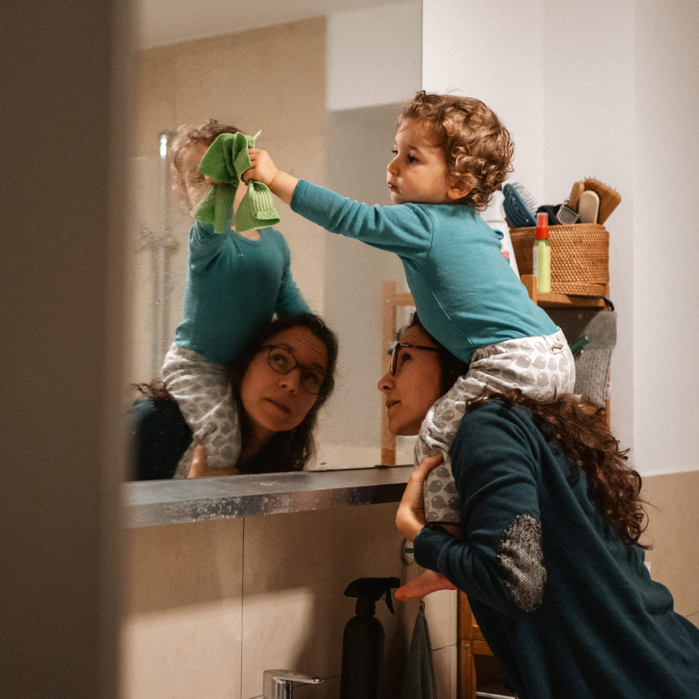Kind sitzt auf den Schultern der Mutter und putzt den Spiegel