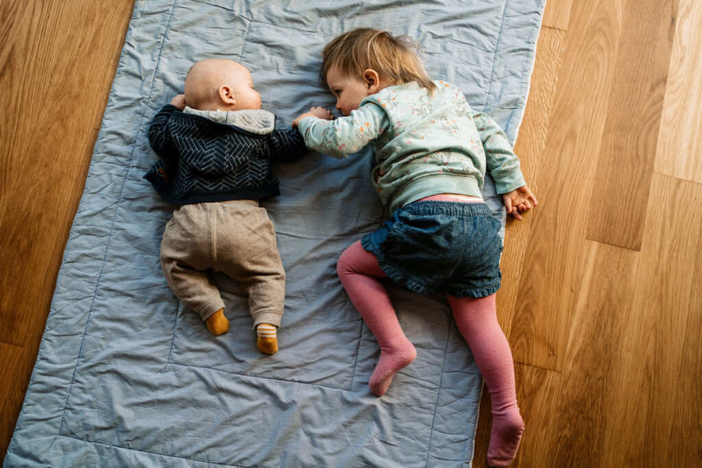 Zwei Geschwister liegen nebeneinander auf einer Decke im Wohnzimmer