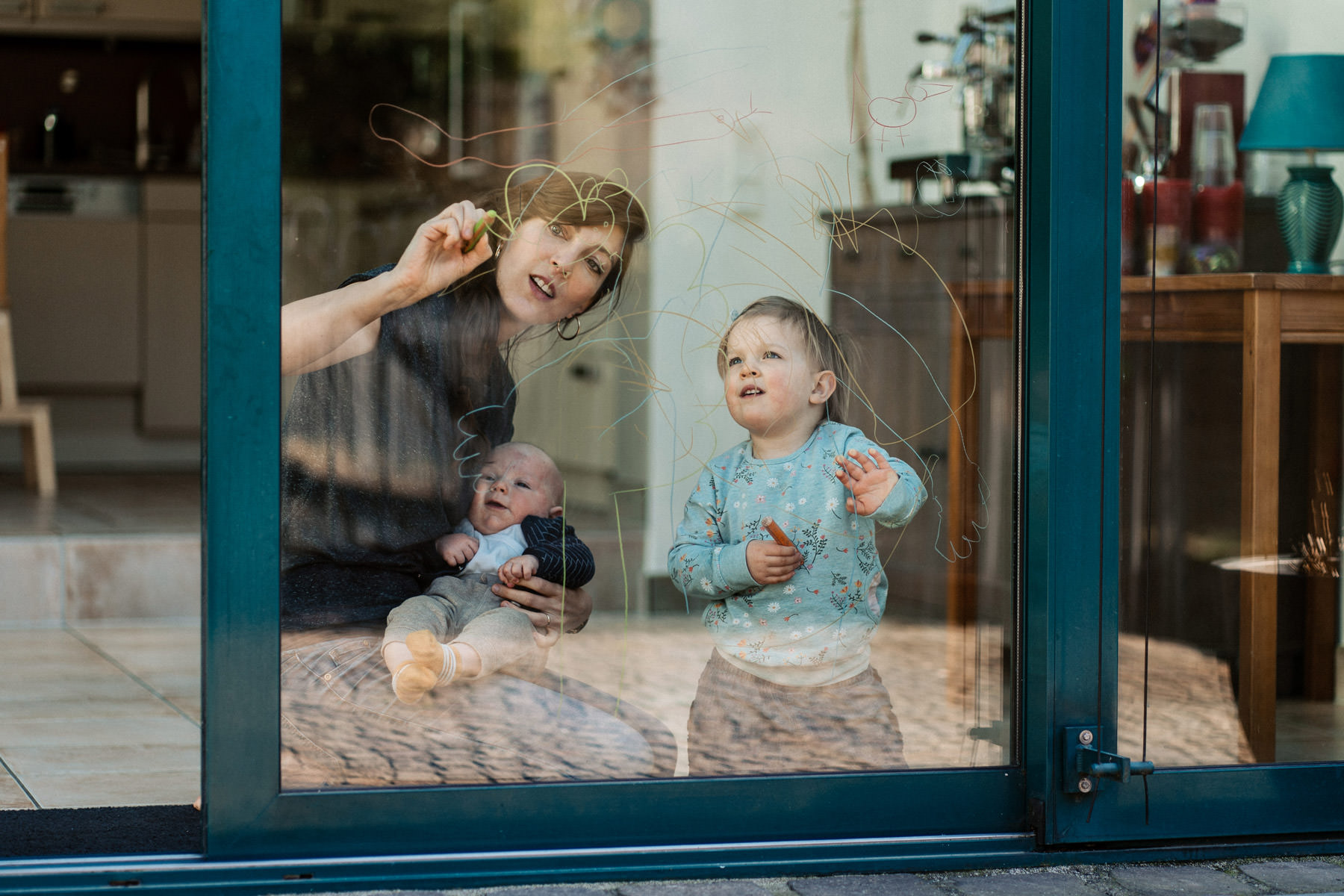 Kinder stehen am Fenster und malen auf die Scheibe, während ein Baby daneben liegt – dokumentarische Familienfotografie in Köln.