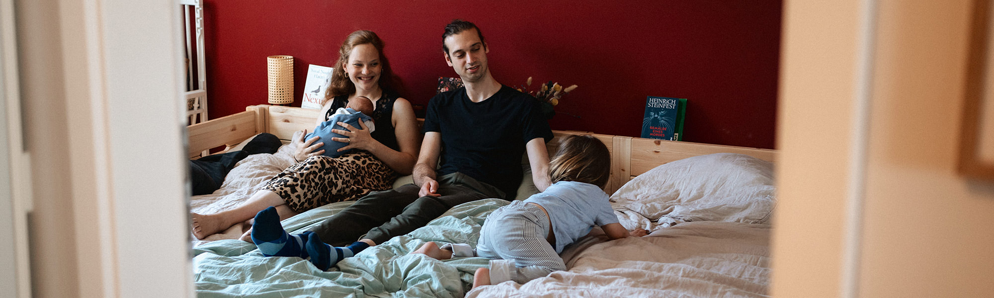 Familie liegt gemeinsam auf einem Bett, fotografiert aus dem Türrahmen.