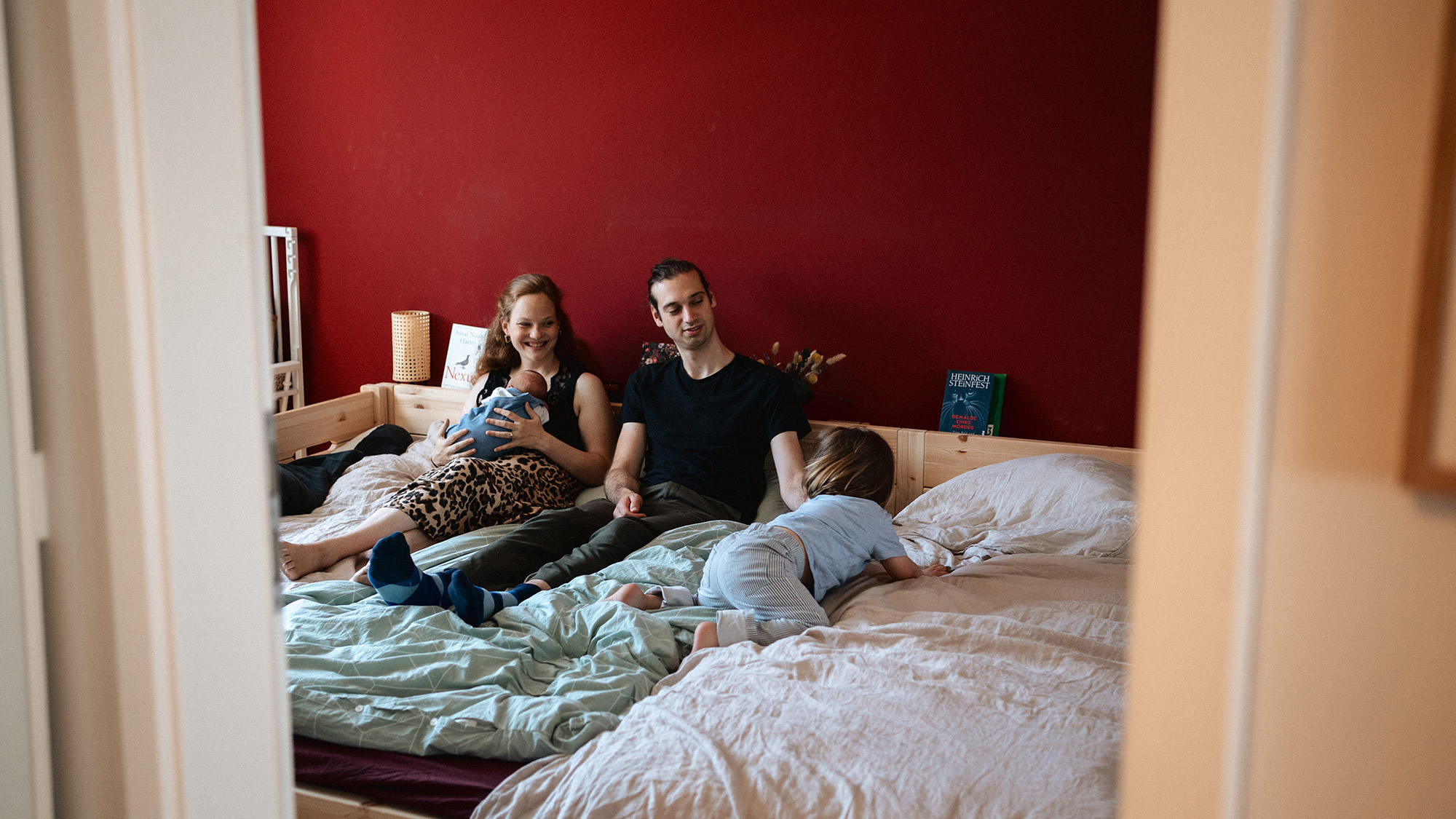 Familie liegt gemeinsam auf einem Bett, fotografiert aus dem Türrahmen.
