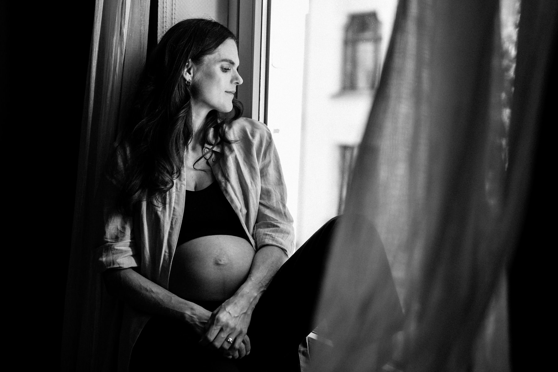 Schwangere Frau sitzt am Fenster und blickt nach draußen, ruhige Schwarzweißaufnahme