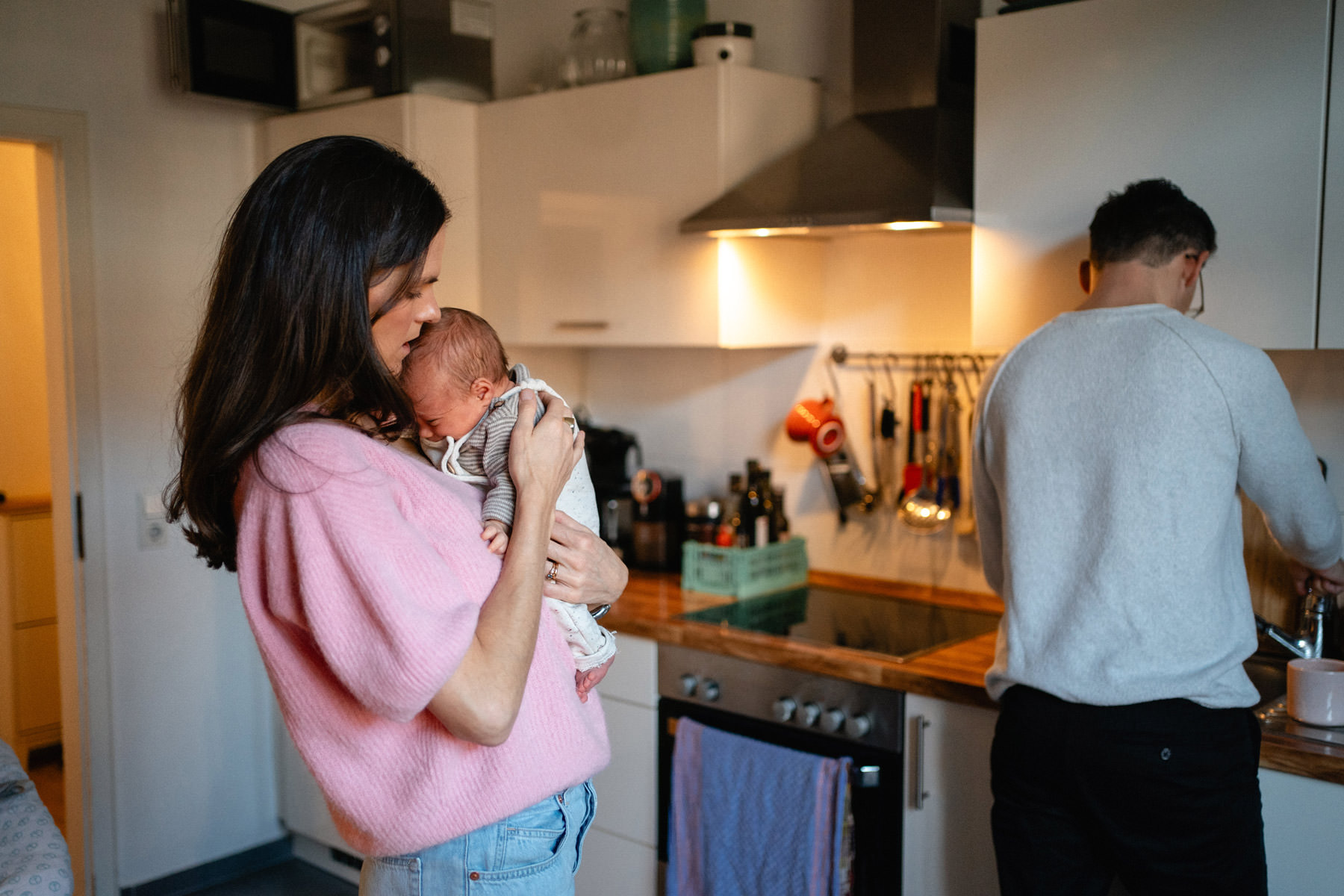 Mama hält ihr Neugeborenes in der Küche, während der Vater im Hintergrund den Alltag übernimmt – Wochenbettfotografie in Köln.