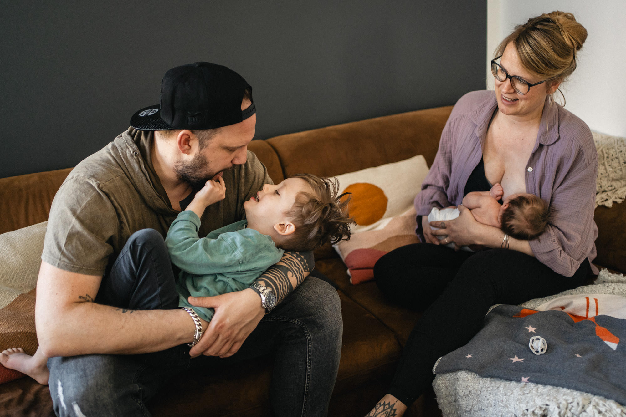 Familie sitzt auf dem Sofa. Die Mutter stillt das Baby, der Papa macht Quatsch mit dem Kleinkind