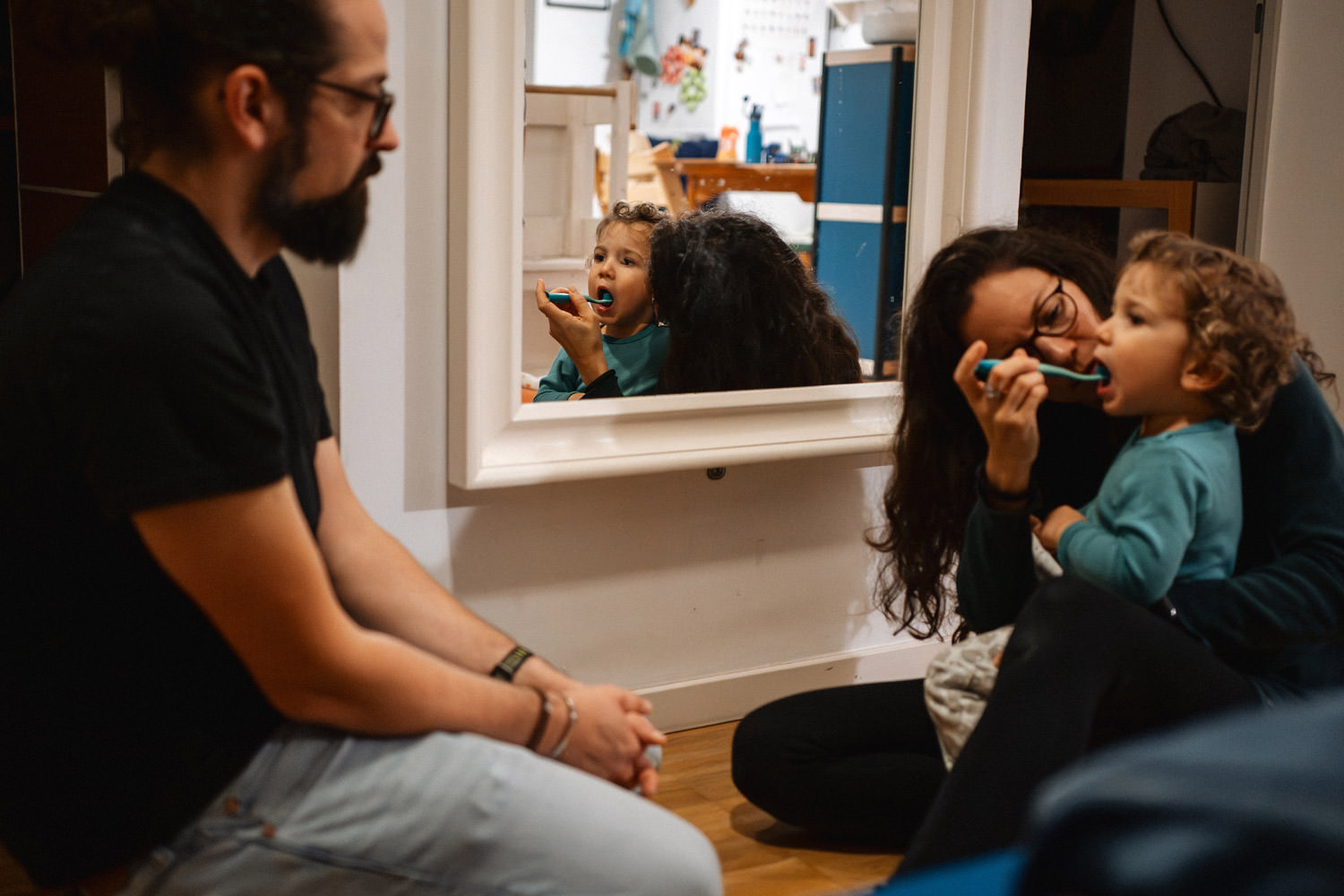 Mutter sitzt auf dem Boden und putzt Kind die Zähne, es spiegelt sich im Wandspiegel und der Vater sitzt daneben