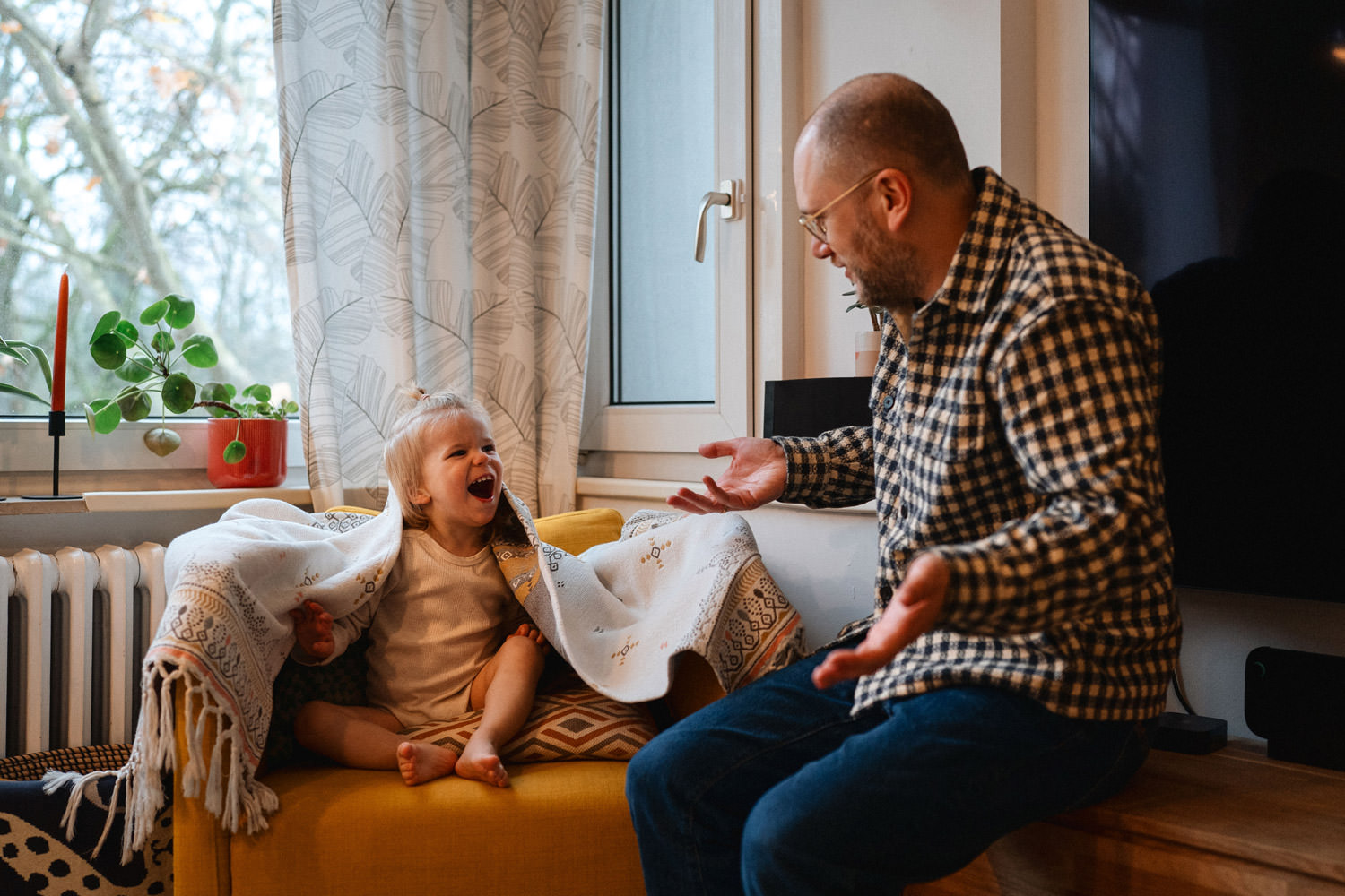 Kind sitzt auf dem Sofa unter einer Decke und lacht den Vater an. Es hat sich versteckt davor