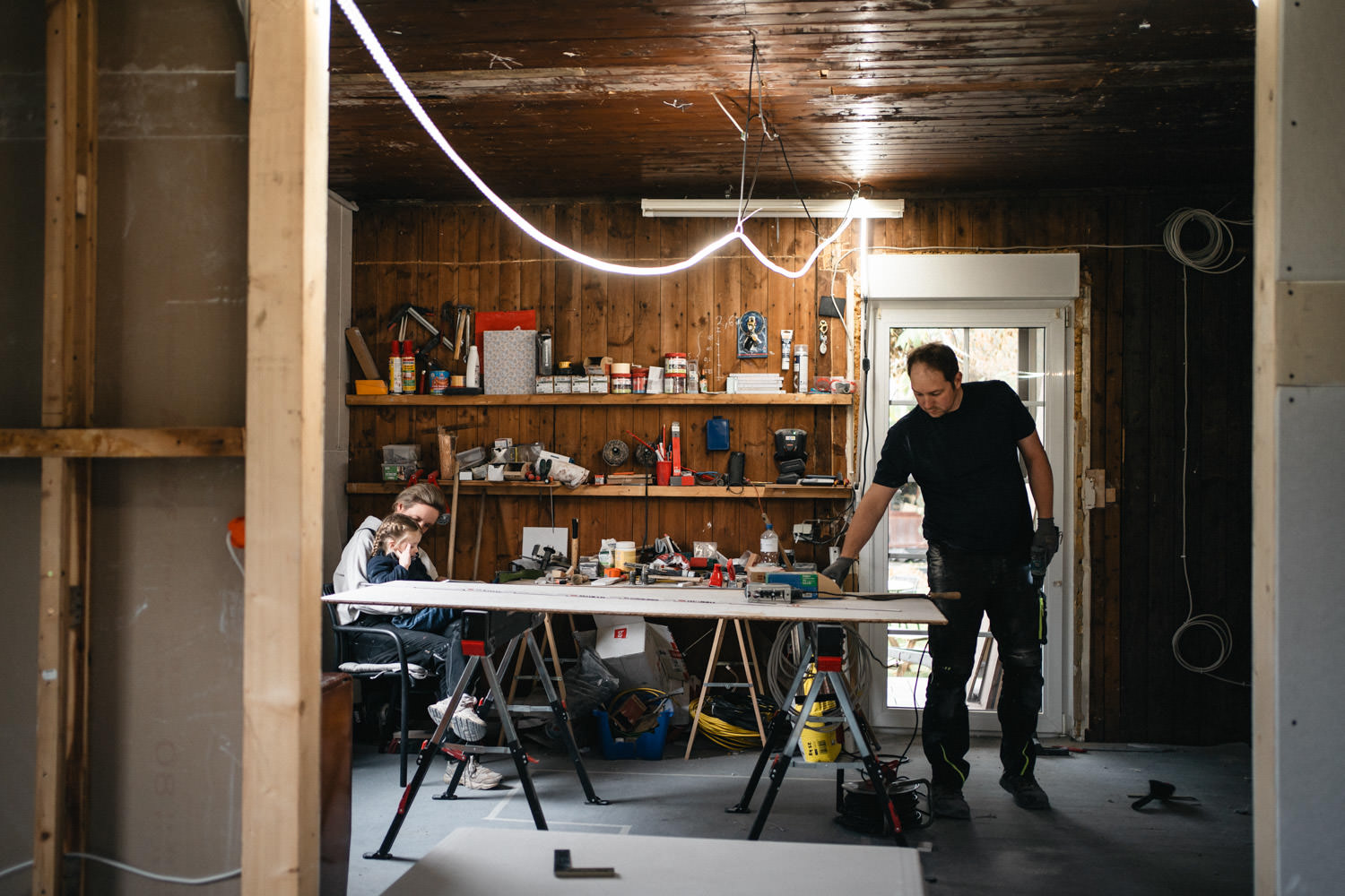 Vater arbeitet in der Baustelle vom Haus, Mutter liest Kind auf dem Schoß etwas vor